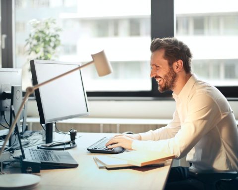 man in white dress shirt sitting on black rolling chair while facing black computer set and smiling