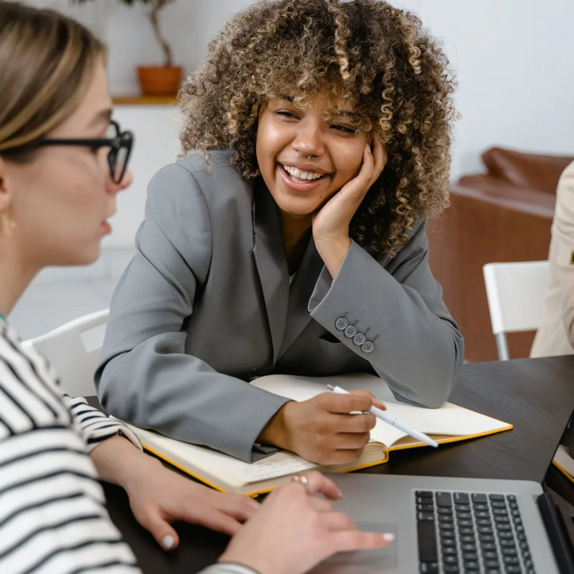 Vrouw in grijze blazer kijkt glimlachend mee naar het werk van een collega achter een laptop op kantoor